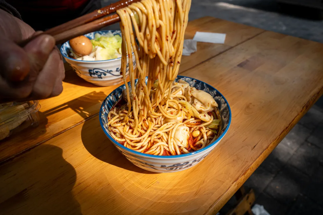 A bowl of spicy Chongqing xiaomian noodles being lifted with chopsticks